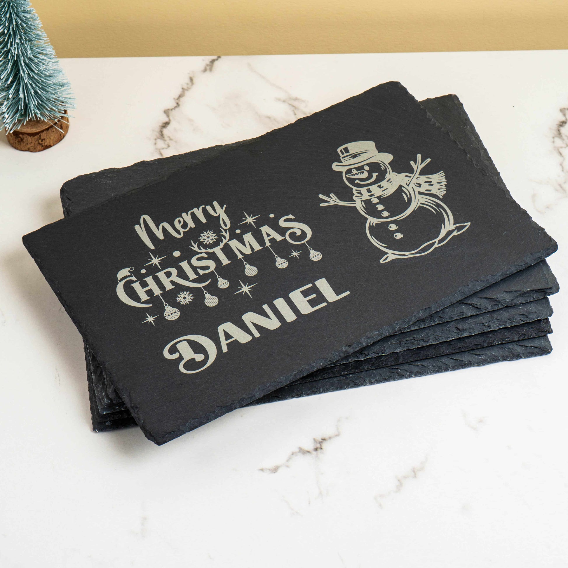 A set of personalised Christmas slate placemats, showing the engraved name "Daniel," stacked to show their rustic edges.