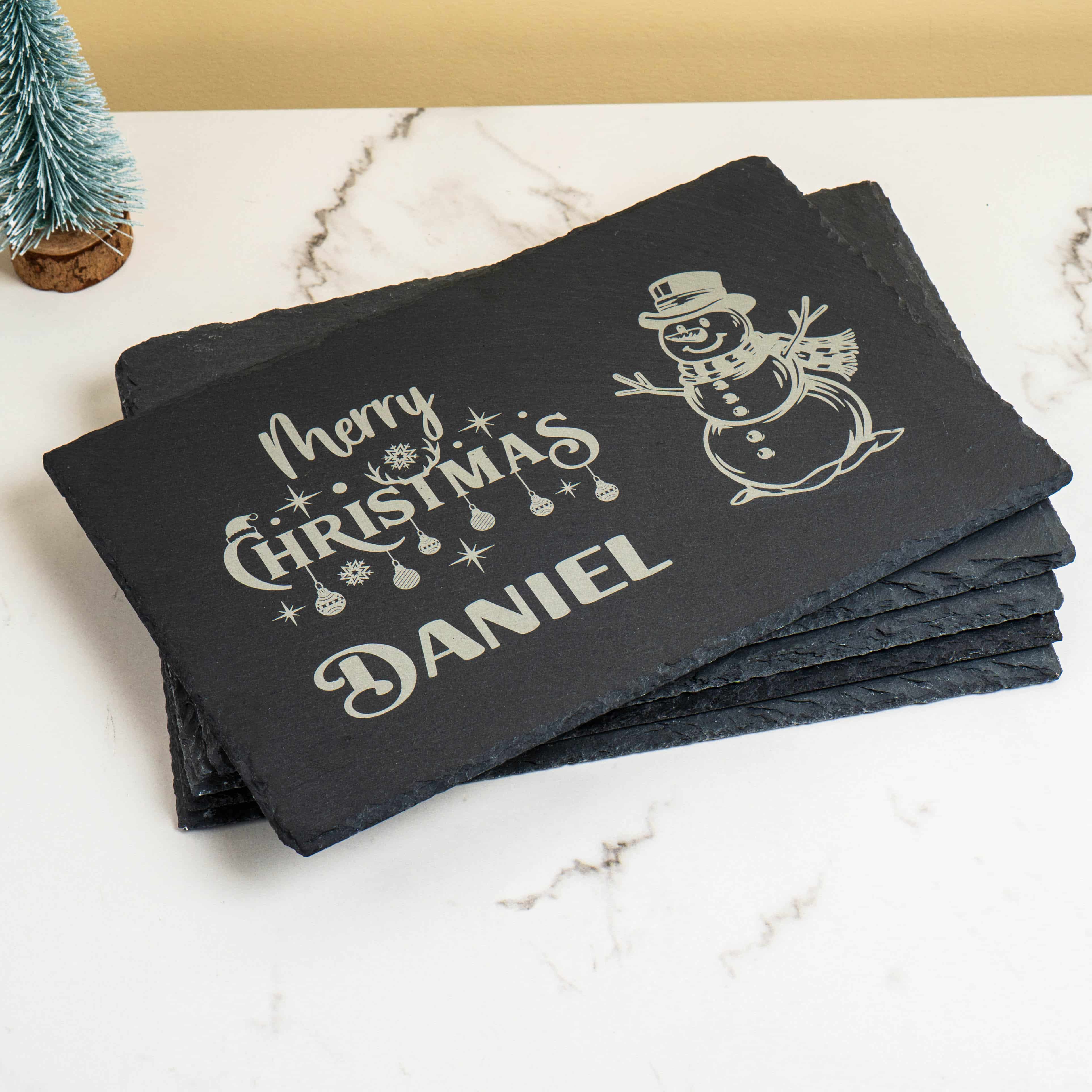A set of personalised Christmas slate placemats, showing the engraved name "Daniel," stacked to show their rustic edges.