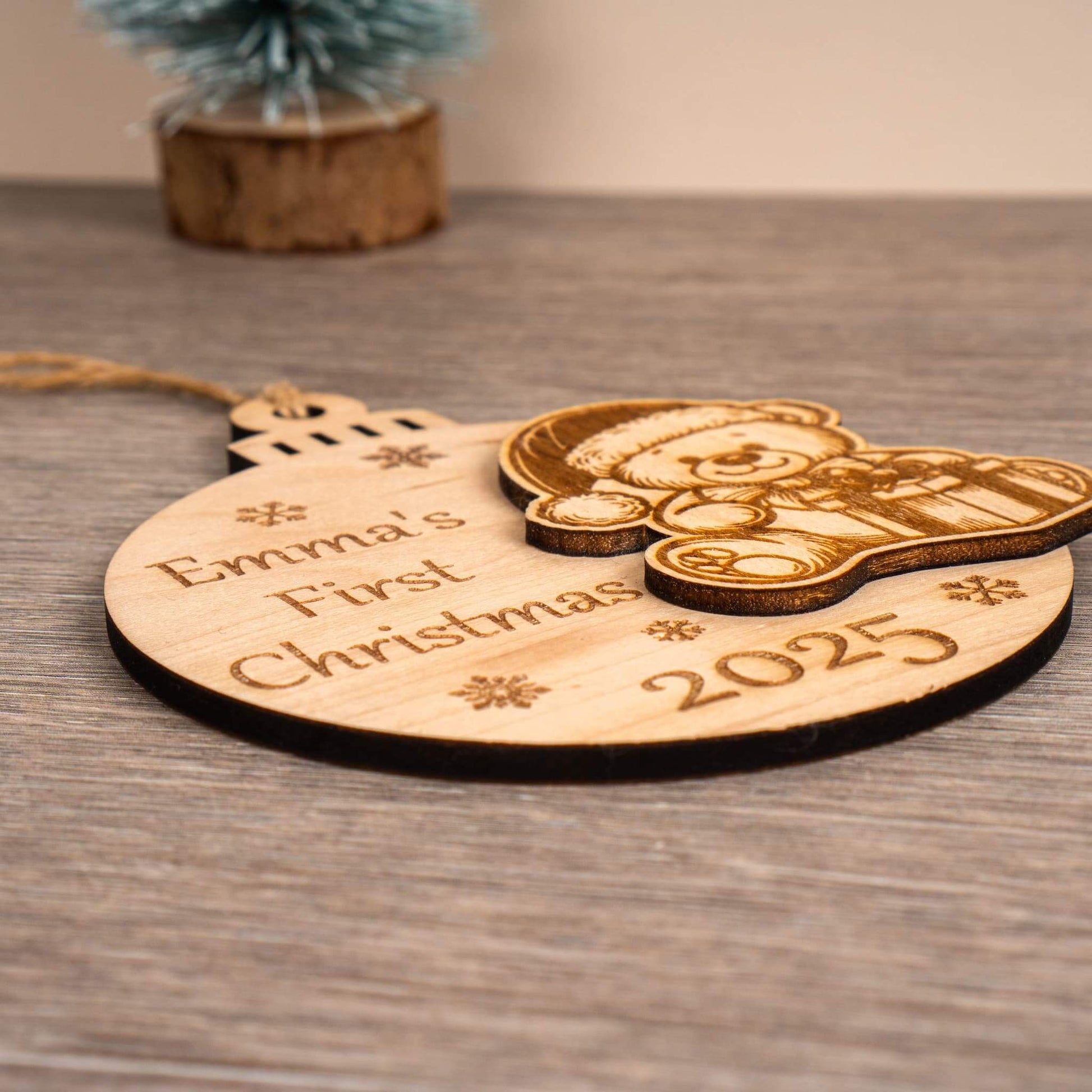 First Christmas bauble with engraved text 'Emma's First Christmas 2025' on a wooden surface.