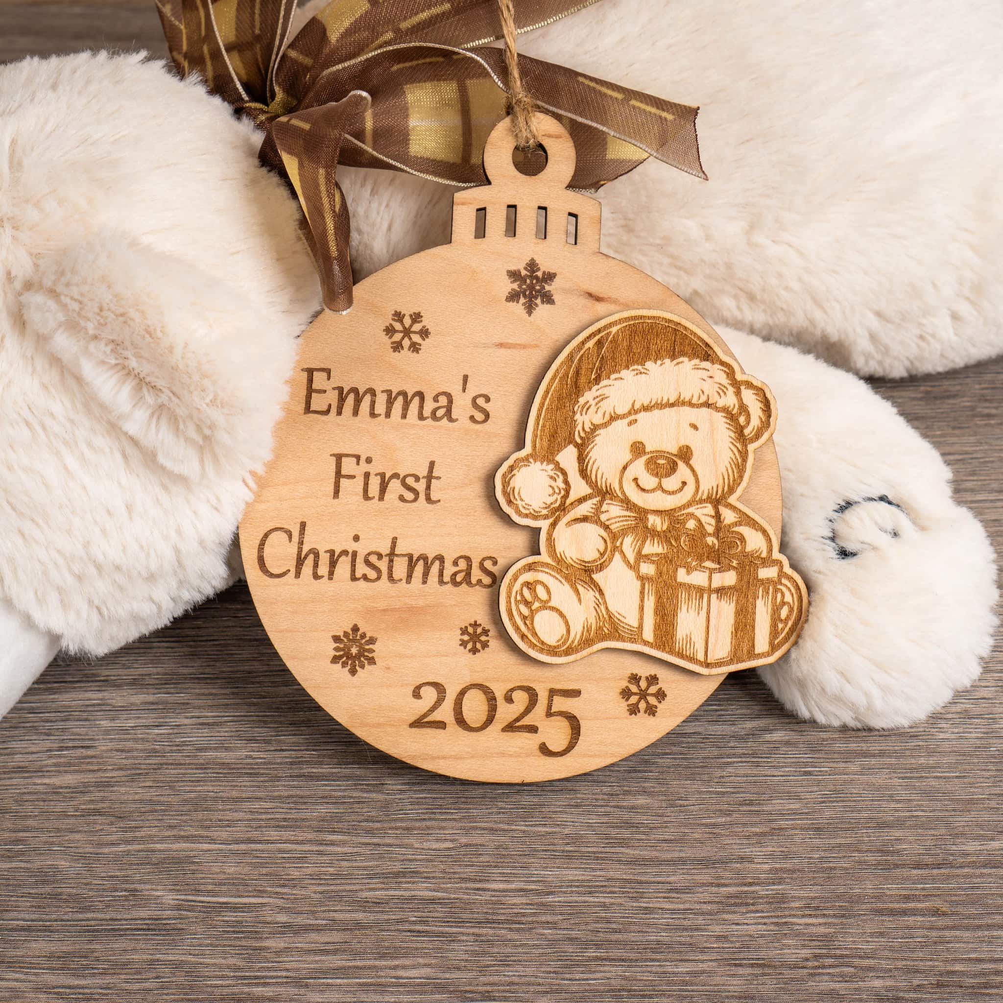 First Christmas Bauble with teddy bear design and text 'Emma's First Christmas 2025' on a wooden table with a soft toy.