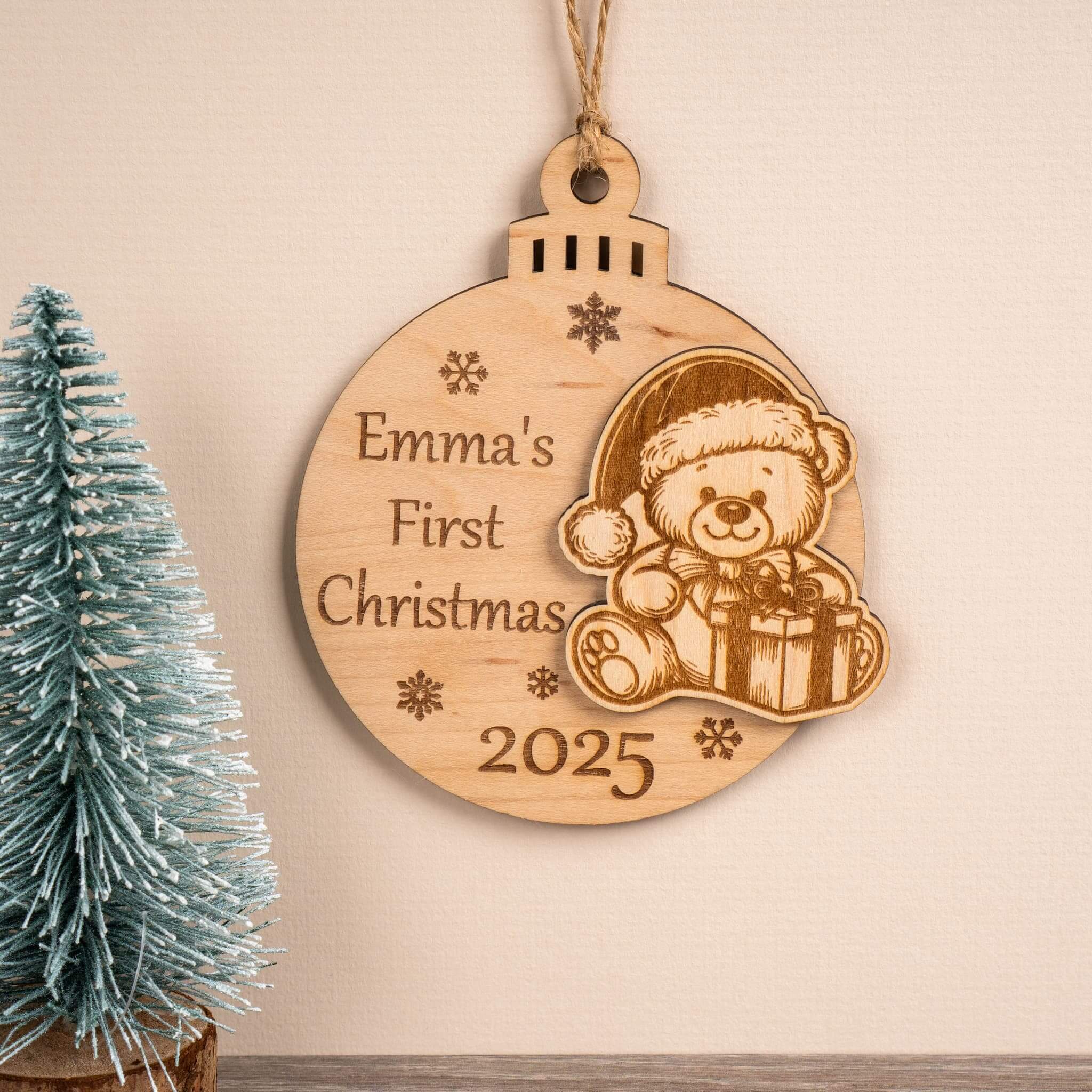 First Christmas bauble with engraved teddy bear and text 'Emma's First Christmas 2025' on a beige background.