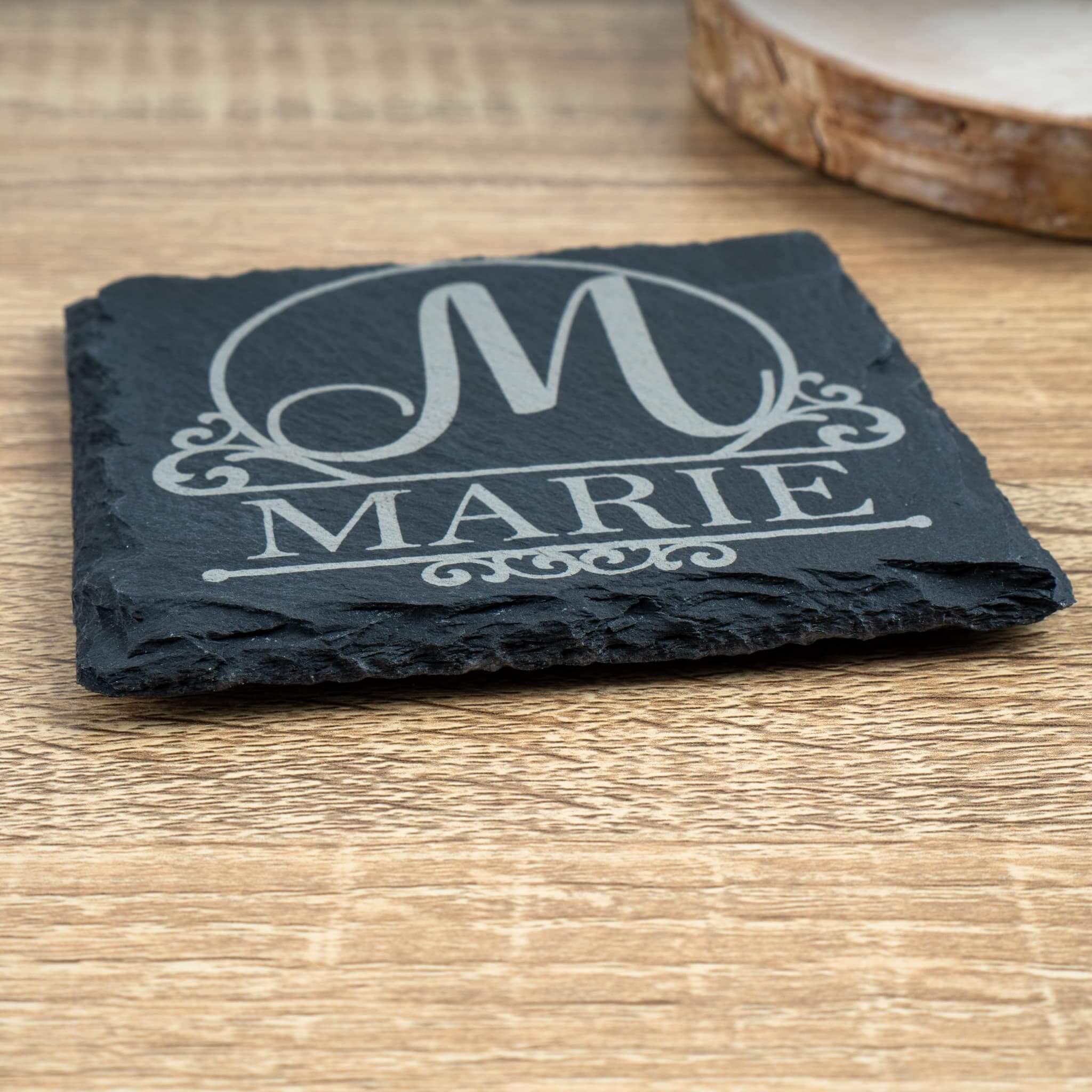 Personalised monogram and name slate coaster engraved with 'M' and 'MARIE', sideview, on rustic wooden table.
