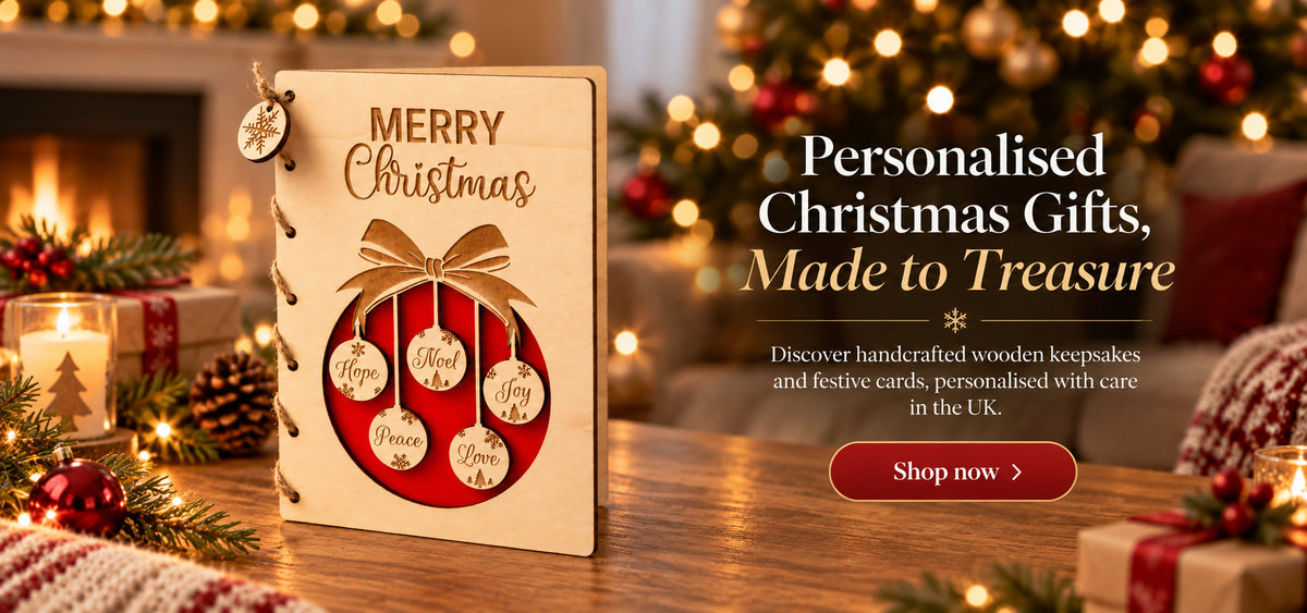 A wooden Christmas greeting card or with 'MERRY Christmas' engraved, featuring a red oval window and hanging ornament cutouts labeled Hope, Peace, Joy, Love, and Noel. The card is styled on a wooden table with Christmas trees, ornaments, and wrapped gifts in the background.
