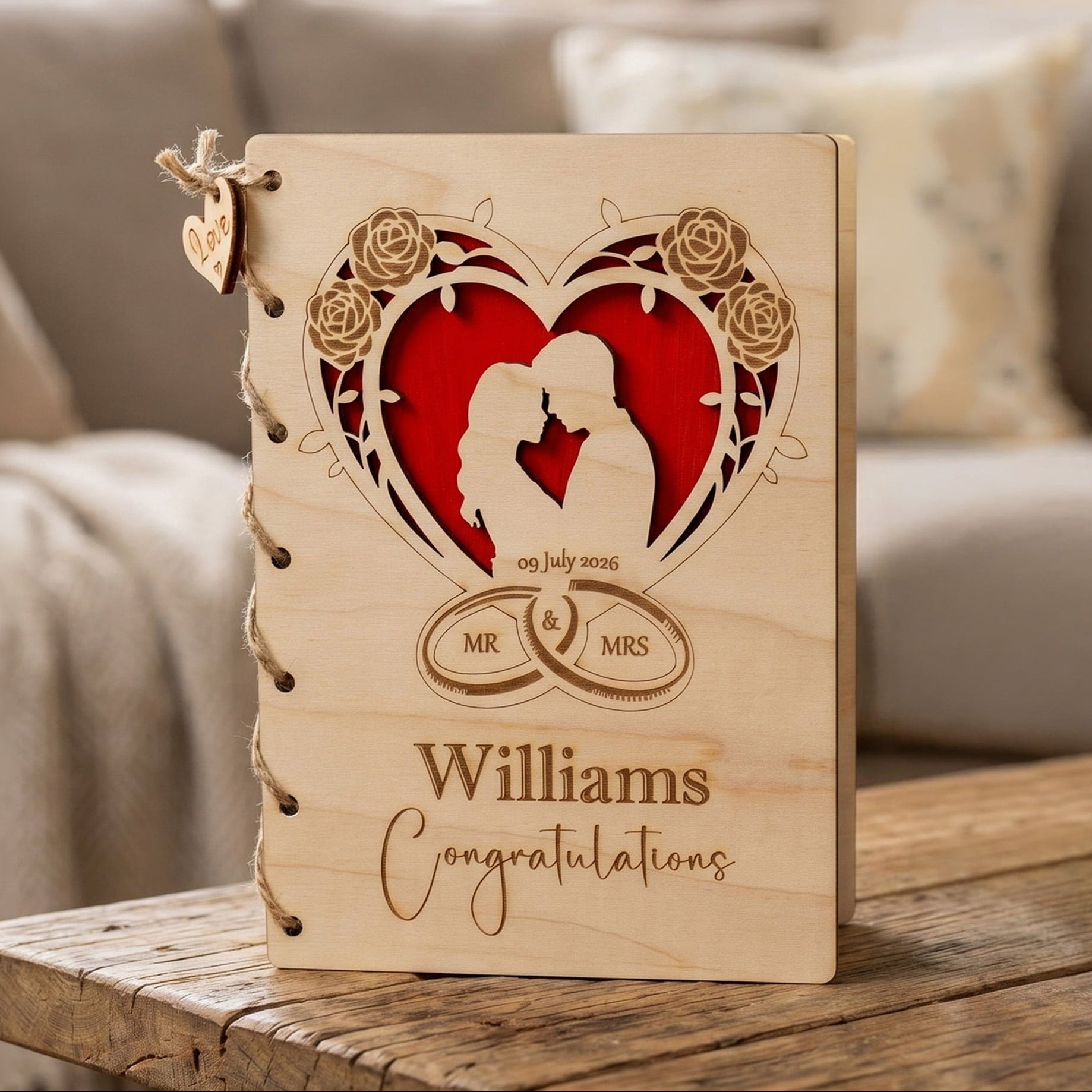 A personalized wooden wedding card, standing on a rustic wooden table in a cozy living room setting. The cover features a laser-engraved heart design with a silhouette of a couple, surrounded by roses, with a red heart insert.