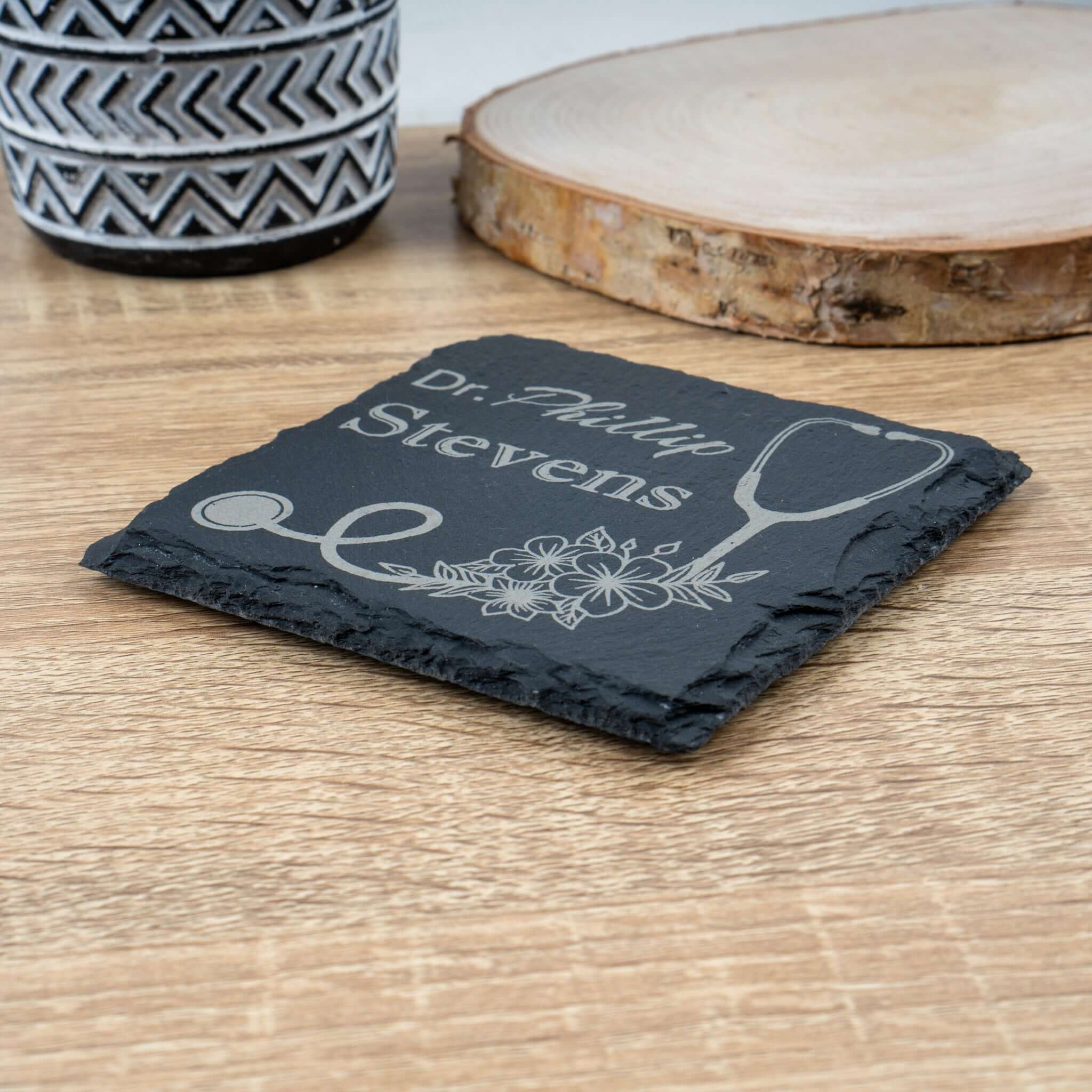 Slate coaster with engraved name and stethoscope design on a wooden surface, side photo.
