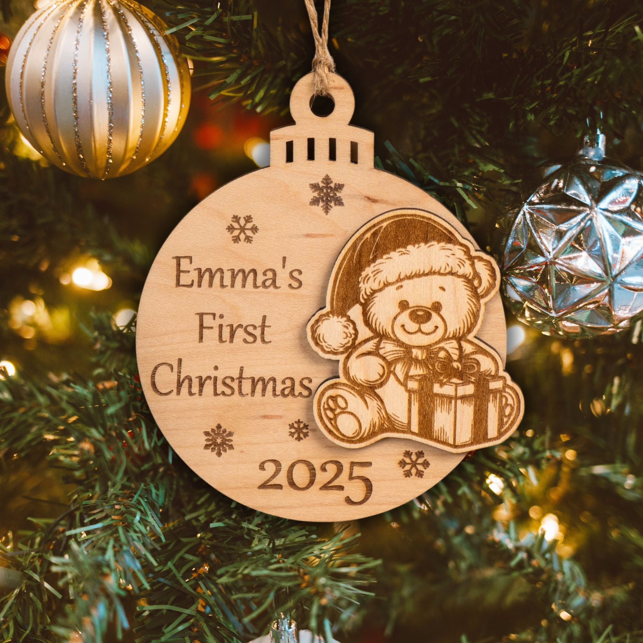 Personalised wooden first Christmas bauble with "Emma's First Christmas 2025" and teddy bear engraving, hanging on a tree.