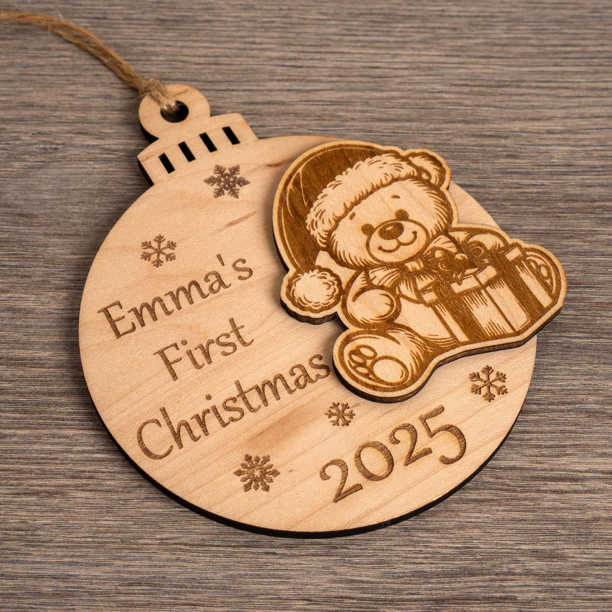 Personalised first Christmas bauble with teddy bear and text 'Emma's First Christmas 2025' on a wooden surface